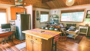 A rustic cabin interior with a wooden ceiling, a central island with a checkered tablecloth, a kitchen area with a refrigerator, and a seating area with a sofa and chairs.