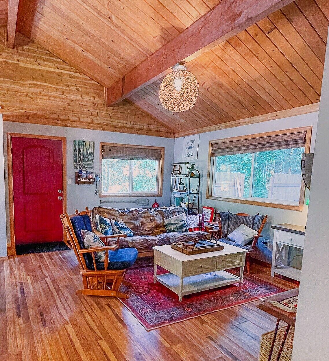 A cozy living room with wooden floors, a red door, a rocking chair, a sofa, a coffee table, and a hanging light fixture.