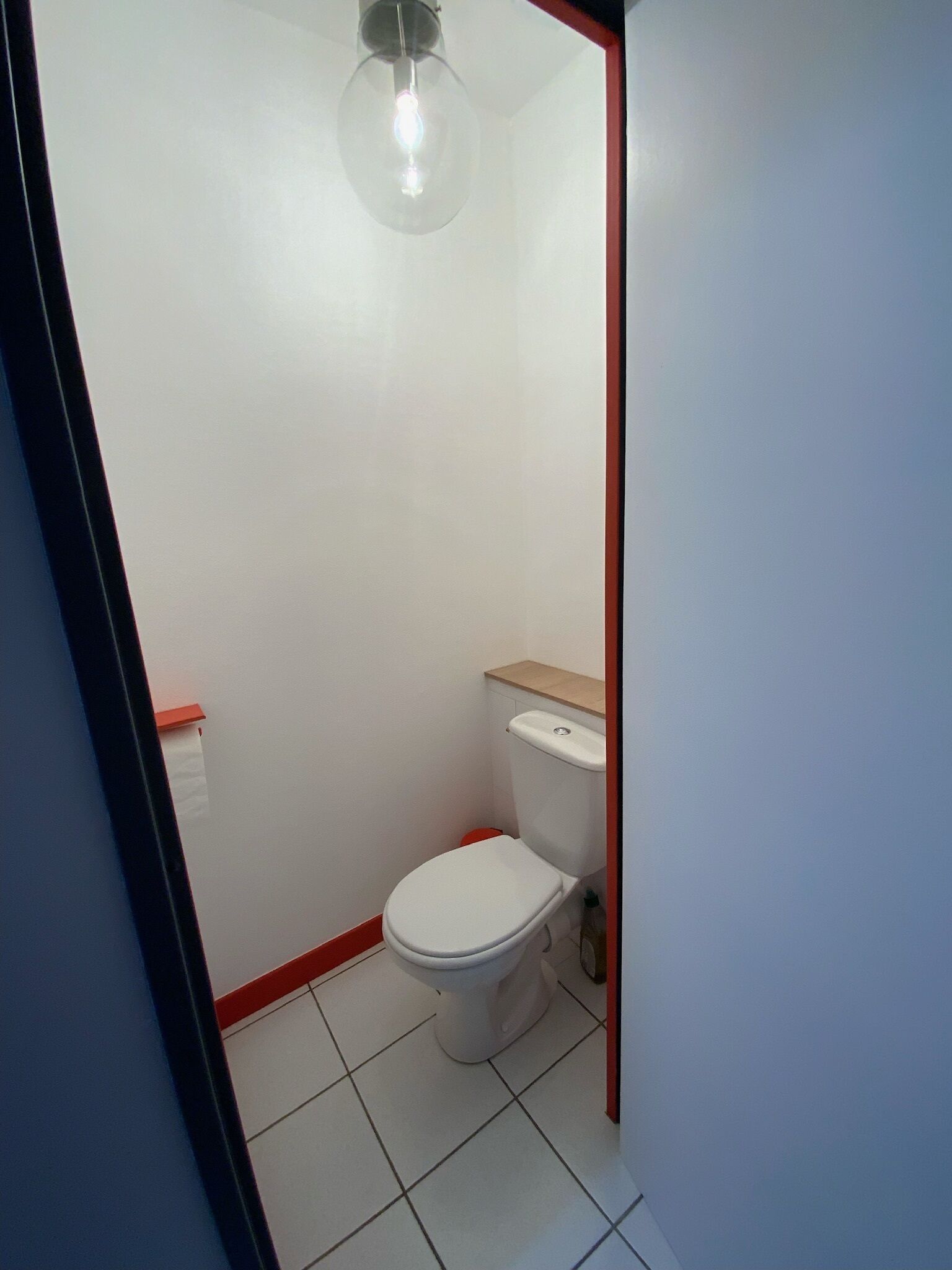 A small, compact bathroom with a white toilet, a wooden shelf above the toilet, and a single light fixture on the ceiling.