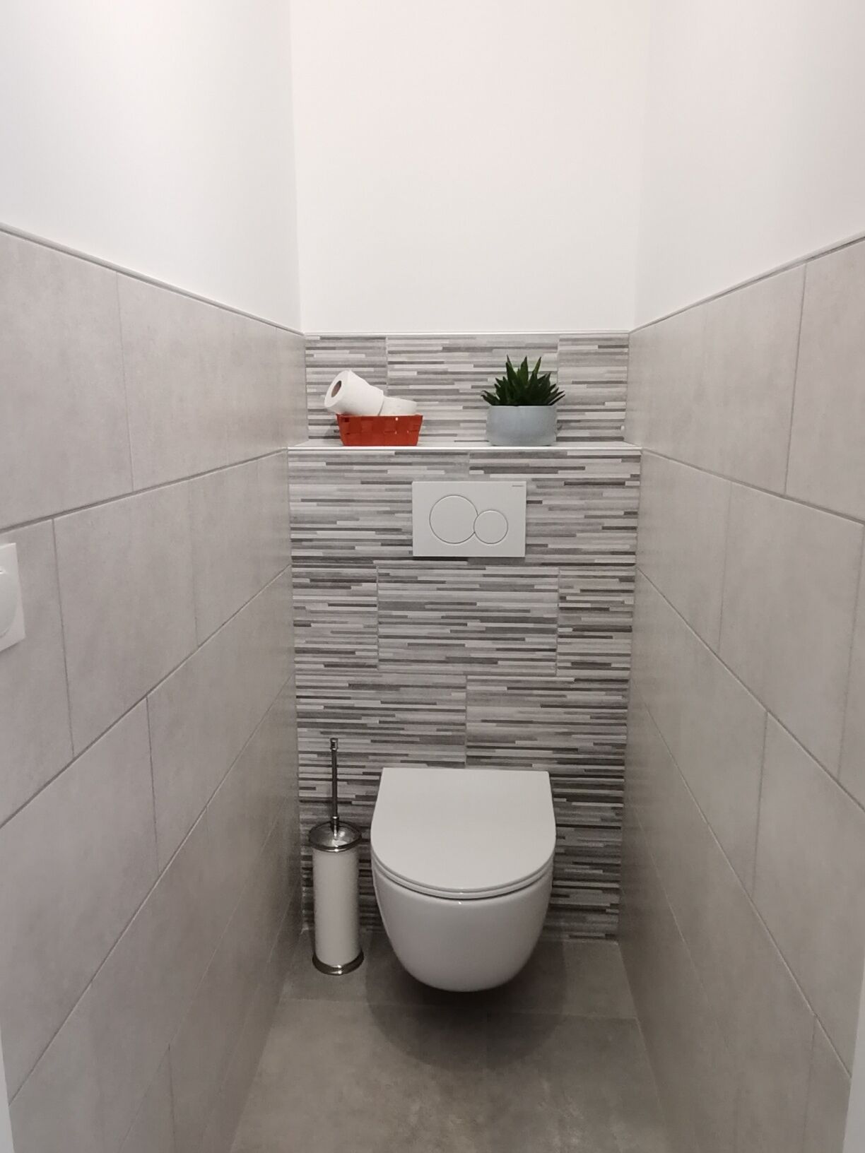 Ein modernes Badezimmer mit Toilette, einem Mülleimer, einer kleinen Ablage mit Pflanze und einer Toilettenpapierrolle.