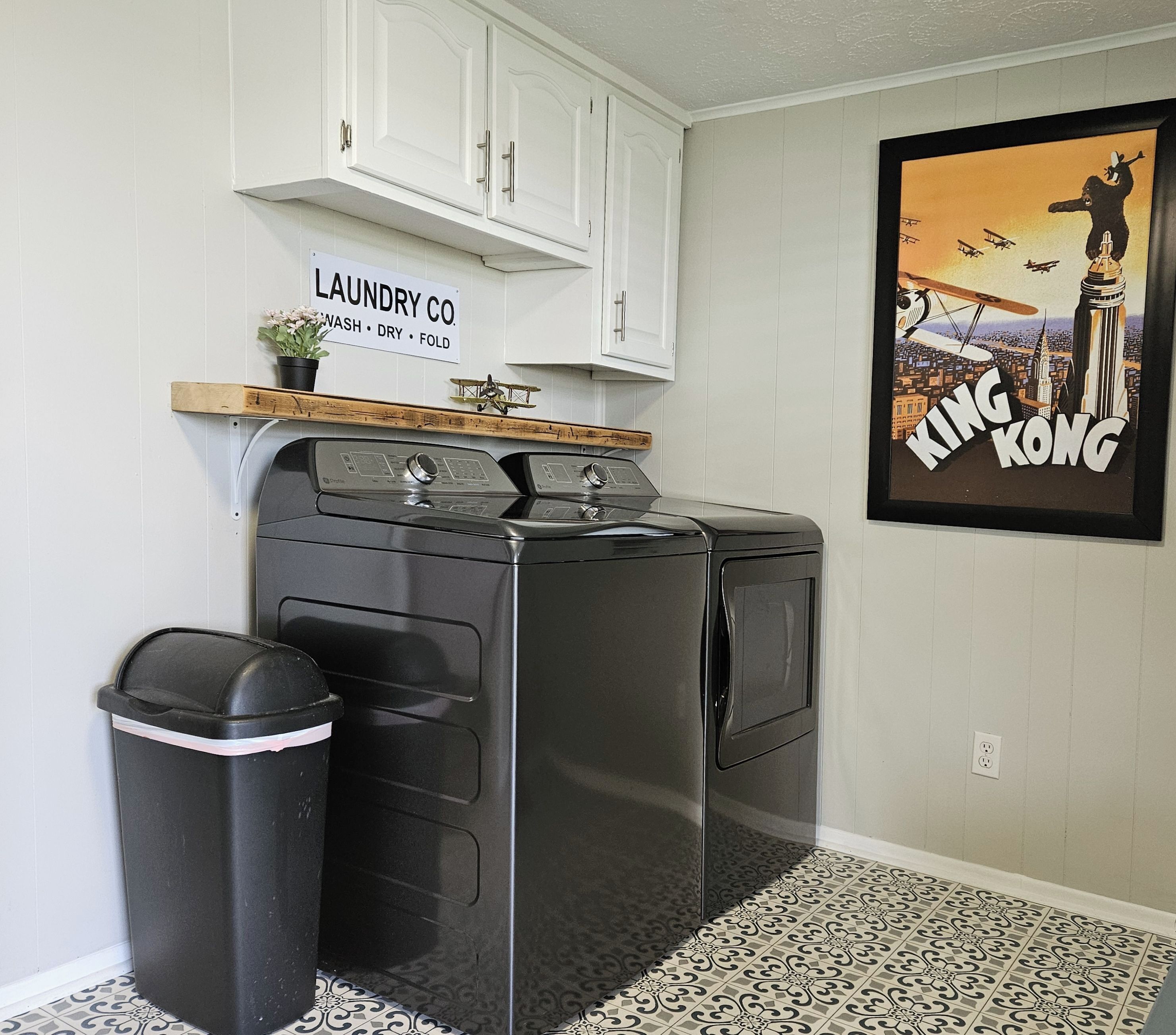 A laundry room with a front-loading washer and dryer, a trash can, and a sign above the washer that reads "LAUNDRY CO. WASH DRY FOLD".
