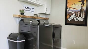 A laundry room with a front-loading washer and dryer, a trash can, and a sign above the washer that reads "LAUNDRY CO. WASH DRY FOLD".