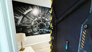 Una habitación con una caja fuerte negra grande, un escritorio blanco con lámpara y un mural en la pared con temática espacial.