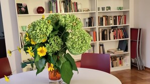 A round table with a vase of flowers in the center, surrounded by red chairs. Behind the table is a white bookshelf filled with books and decorative items. - Spacious Living in a small 50s villa with large garden and fireplace (Langen (Hessen))
