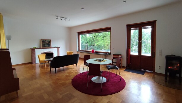 A spacious living room with a fireplace, a dining area with a round table, and large windows offering a view of greenery. - Spacious Living in a small 50s villa with large garden and fireplace (Langen (Hessen))