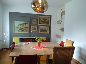 Un coin repas avec une table en bois, des chaises rouges et jaunes, un fauteuil en osier et un luminaire suspendu.