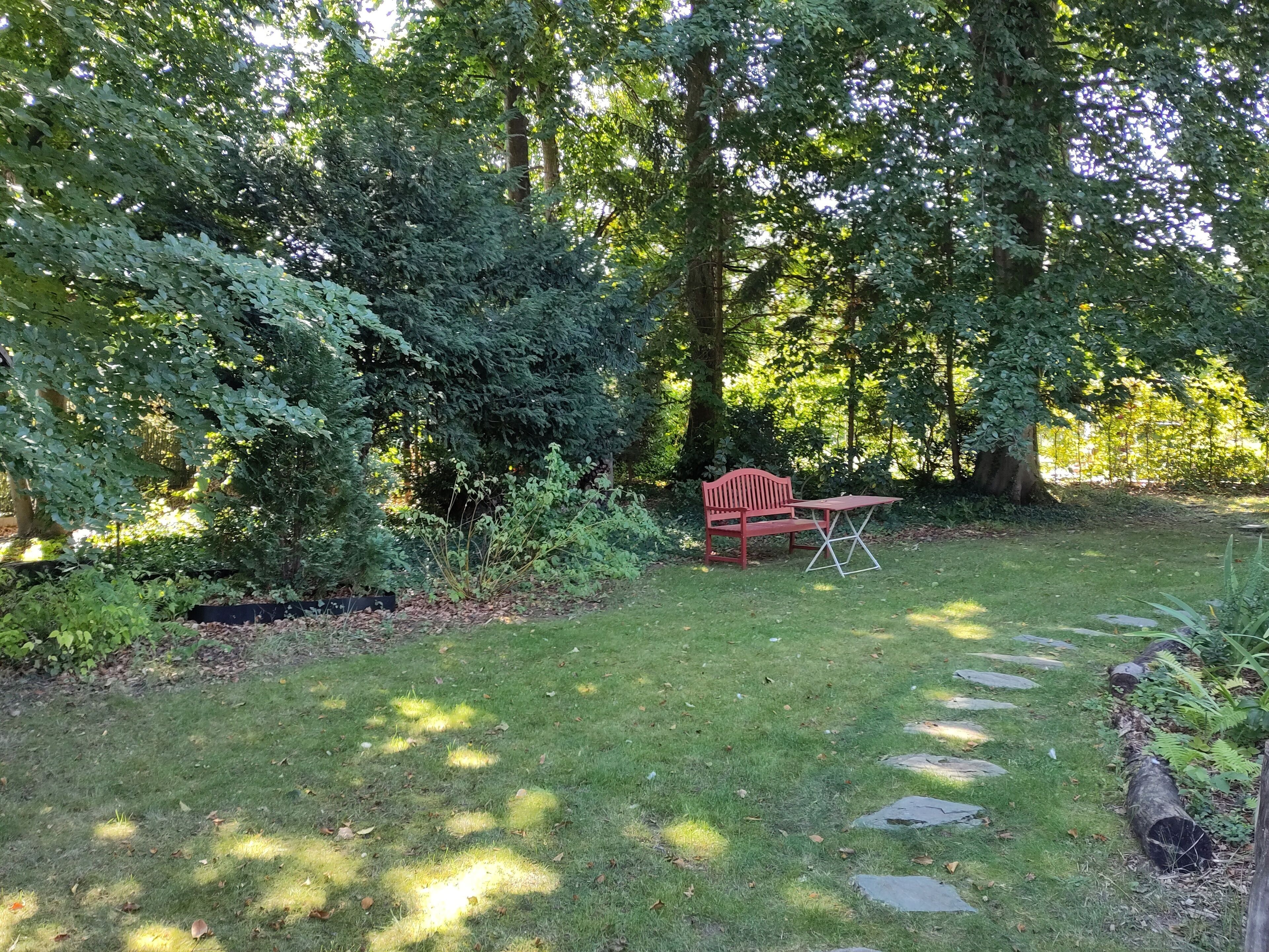 A garden with a red bench and a table, surrounded by trees and a stone pathway.