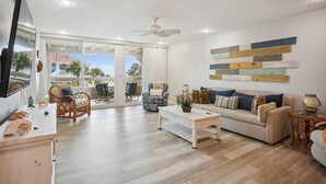 A spacious living room with a sofa, coffee table, and wicker chairs. A ceiling fan is visible, and there is a TV mounted on the wall. - Ocean View Condo in St. Augustine (Saint Augustine)
