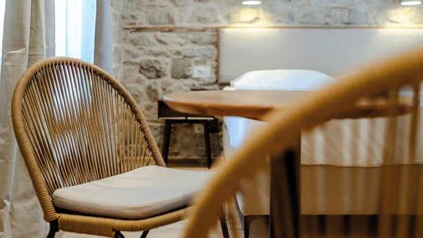 A rustic room with a stone wall, a wooden ceiling, a wicker chair, a wooden table, and a bed with white bedding. - Porto Nativo Split (Split)