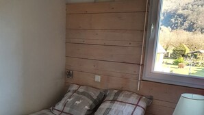 Ein Schlafzimmer mit einem hölzernen Kopfteil, einer karierten Bettdecke, zwei Kissen, einem Nachttisch mit Lampe und einem Fenster mit Blick auf Garten und Hügel.