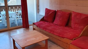 Ein gemütliches Wohnzimmer mit einer Holzsofa, einem kleinen Holztisch und einer Schiebetür zu einem Außenbereich mit weißen Stühlen.