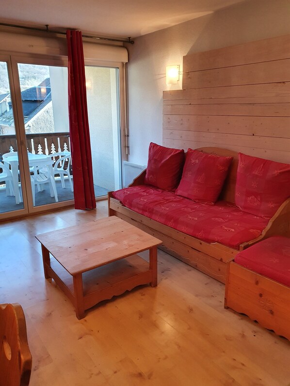 A cozy living room with a wooden sofa, a small wooden table, and a sliding glass door leading to an outdoor area with white chairs.