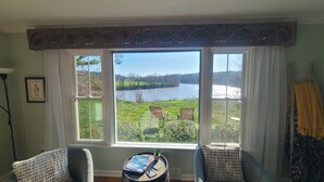 Una habitación con un amplio ventanal que da a un río, dos sillas, una mesita con una revista y una lámpara.