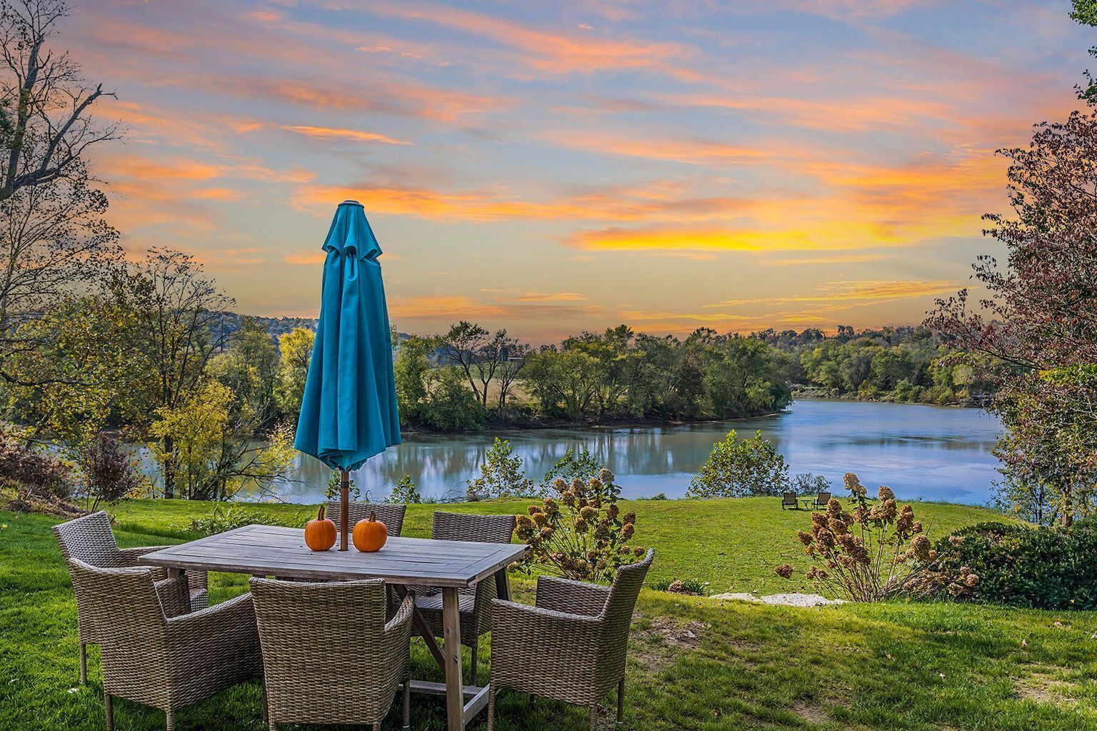 A table with wicker chairs and a blue umbrella overlooking a lake with trees and hills in the background.