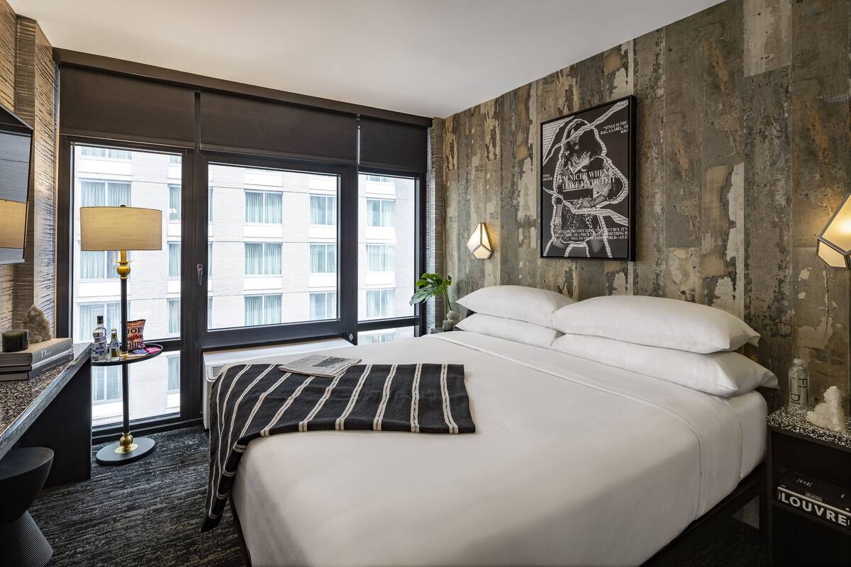 A hotel room with a large bed, a desk with a lamp, a wall-mounted artwork, and a view of the city through large windows.