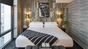 A modern bedroom with a large bed, a black and white striped throw, two bedside lamps, a poster on the wall, and a book titled 'LOUVRE' on the bed.