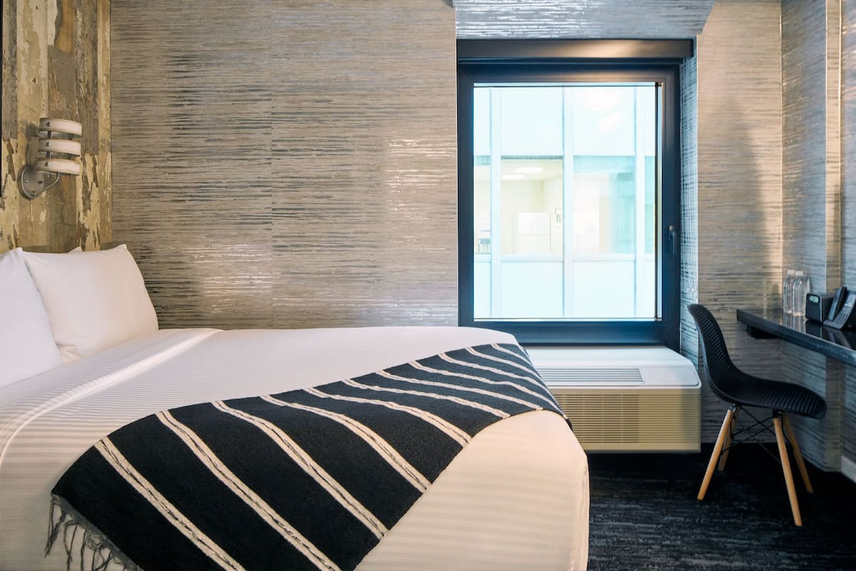 A hotel room with a large bed, a window with blinds, a desk with a chair, and a wall-mounted air conditioning unit.