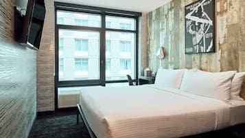A hotel room with a large bed, a flat-screen TV mounted on the wall, a poster on the wall, and a view of the city through large windows.