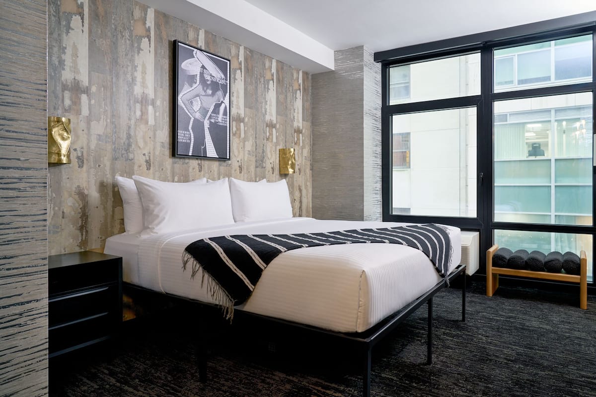 A modern hotel room with a large bed, a wooden headboard, a framed picture on the wall, and a large window offering a view of the city.