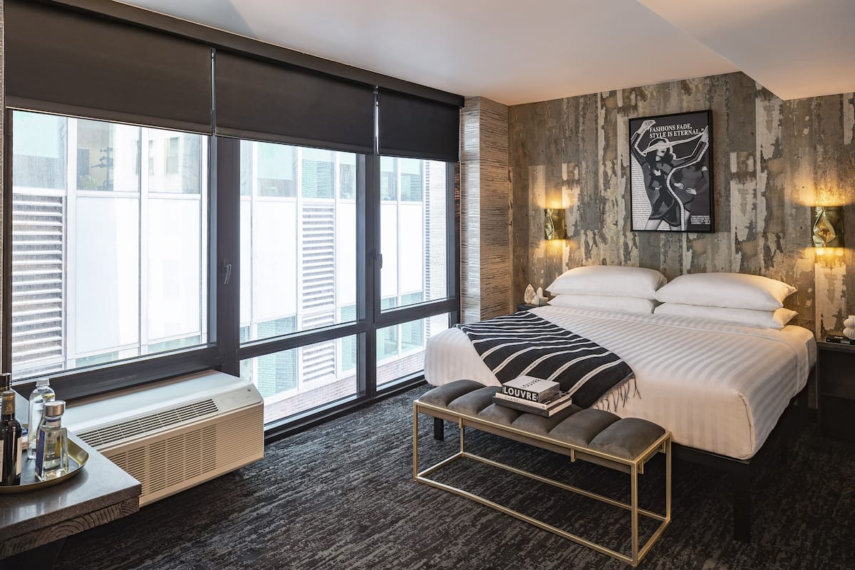 A modern bedroom with a large bed, a bench with a book, a radiator, and a city view through large windows.