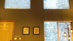 A room with a high ceiling, two large windows with blinds, a door, and a couch with a patterned cushion. - Private Cabin with Open Loft - Sleeps 3-6 (Seward)