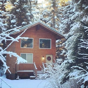 A wooden cabin with a porch and a wreath on the door, surrounded by snow-covered trees.