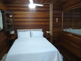 Una habitación con paneles de madera, una cama, un ventilador de techo, una mesita de noche con lámpara y una ventana con vistas.