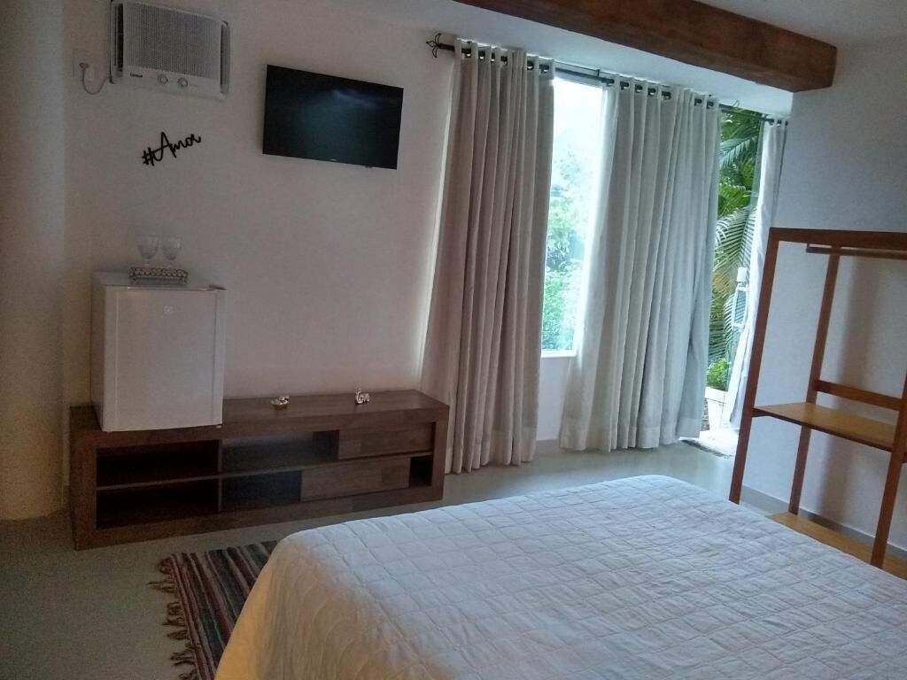 Ein Schlafzimmer mit einem Bett, einem an der Wand montierten Fernseher, einem kleinen Schrank und einem Fenster mit Vorhängen.
