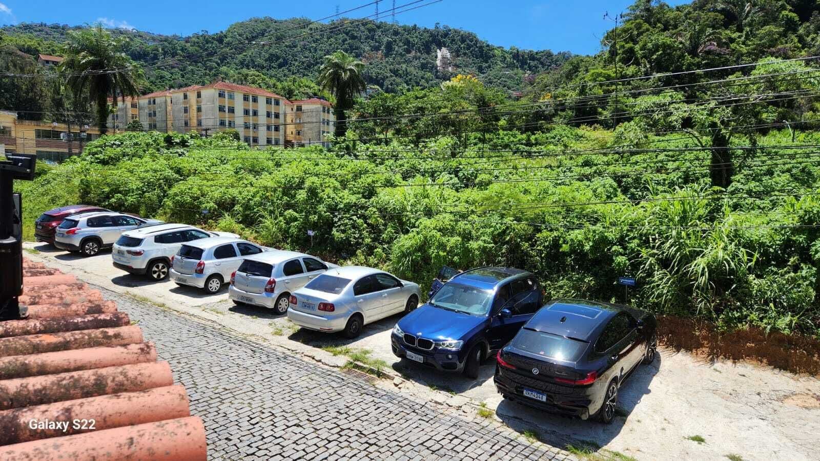 Uma área de estacionamento com vários carros, um edifício ao fundo e um morro com vegetação.