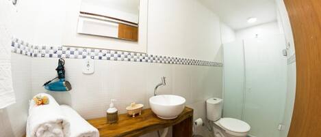 A bathroom with a wooden vanity, white sink, and a mirror above it. There is a towel rack with towels, a soap dispenser, and a small bottle on the vanity. A toilet and a shower are visible in the background.