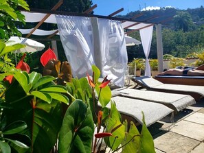 Zona junto a la piscina con sillones de descanso, una pérgola con tela blanca y abundante vegetación.
