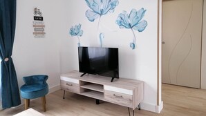 Ein Zimmer mit Fernseher auf einer Holzkommode, einem blauen Sessel und einer Wand mit floralem Wandbild.