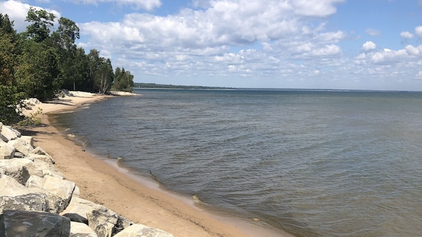 A beach with large rocks in the foreground, a sandy shore, and a calm sea under a partly cloudy sky. - Pure Michigan on Lake Michigan-Manistique, miles of sandy beach fun (Manistique)