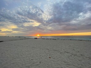 A beach with a sunset in the background, sandy shore, and a few people walking.