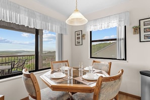 Un coin repas avec une table dressée pour quatre personnes, de grandes fenêtres avec des rideaux et une vue sur un paysage.