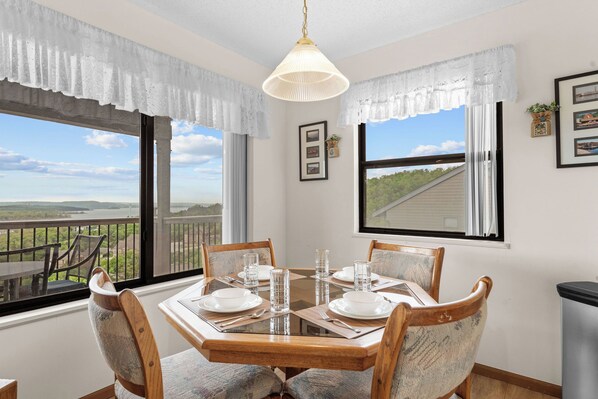 A dining area with a table set for four, large windows with curtains, and a view of a landscape.