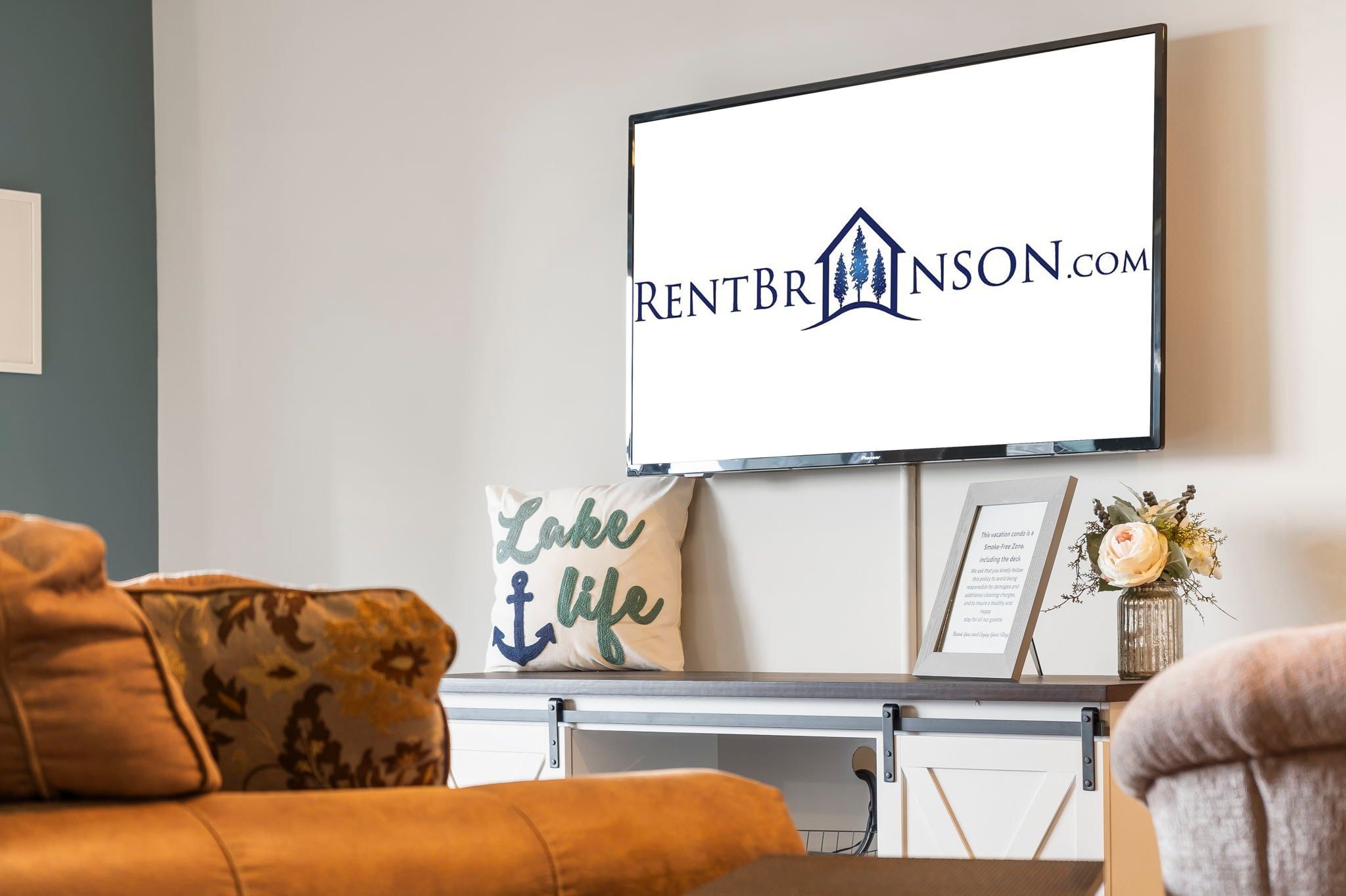 A living room with a mounted television displaying a logo, a sofa with patterned cushions, and a decorative item on a side table.