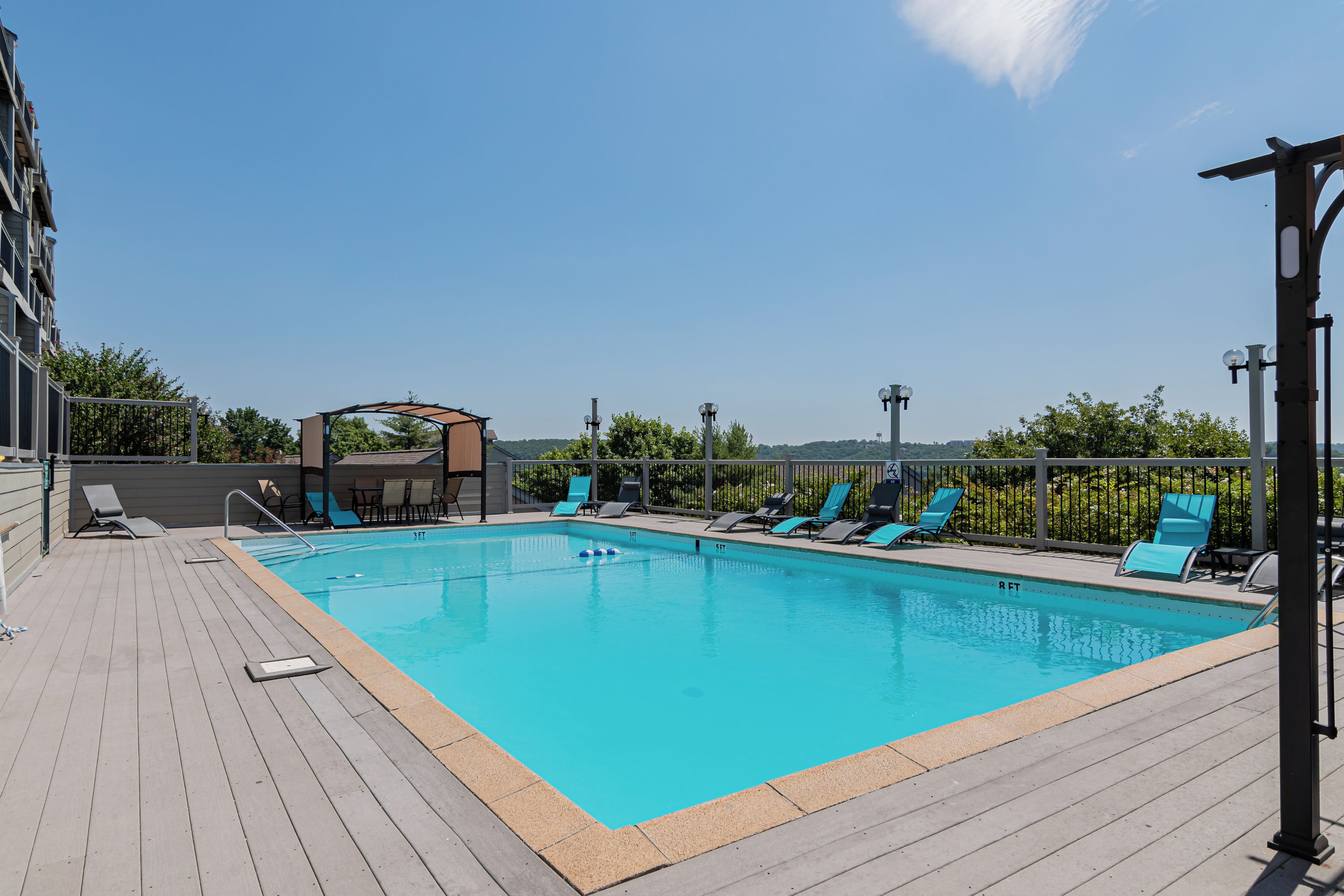 A rectangular swimming pool with a wooden deck, surrounded by blue lounge chairs and a covered seating area.