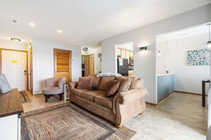 A living room with a brown sofa, a chair, a rug, and a kitchen area visible through an open door.