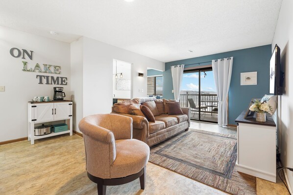 A living room with a sofa, armchair, and coffee table, a TV, and a view of the outdoors.