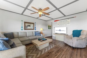 Un salon équipé d’un ventilateur de plafond, d’un canapé, d’un fauteuil, d’une table basse, d’un téléviseur et d’un panneau affichant des conseils.