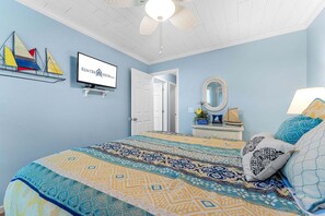 A bedroom with a bed, a television, a ceiling fan, a door, a mirror, and a small shelf with decorative items.
