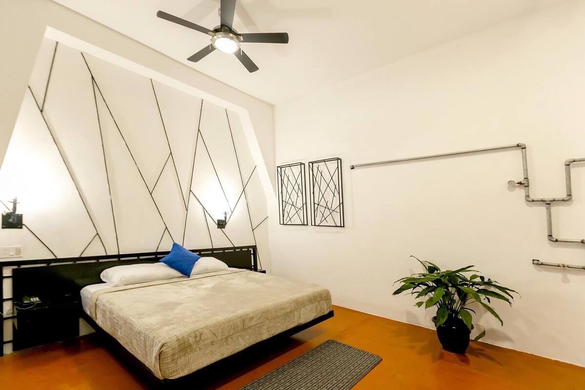 A bedroom with a bed, a ceiling fan, a potted plant, and abstract wall art.