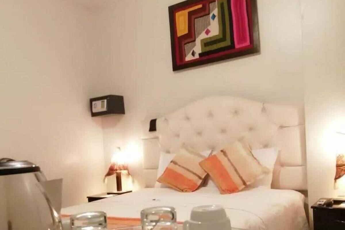 A hotel room with a bed, bedside tables, a glass table with a coffee pot, cups, and a box of chocolates.