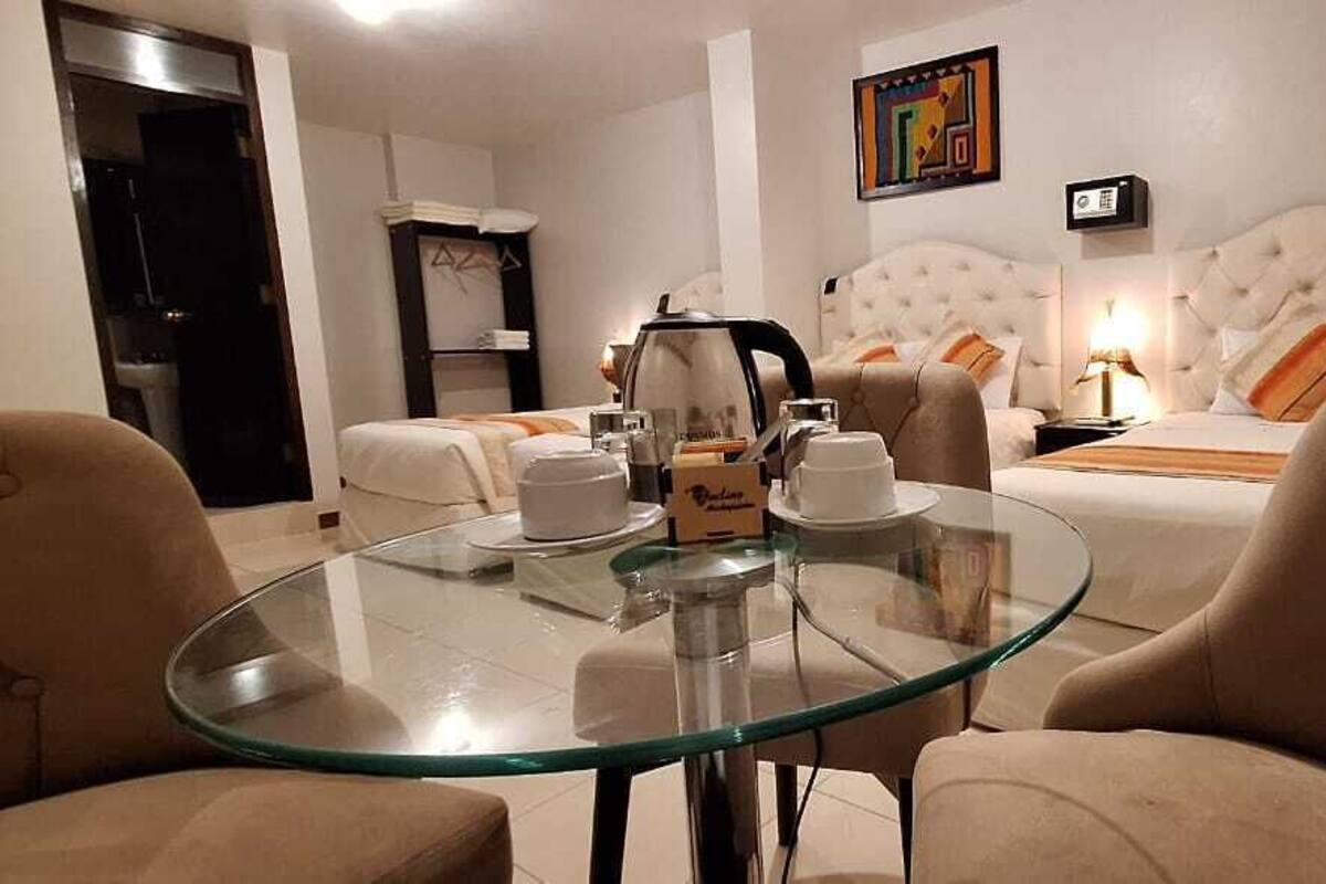 A hotel room with a glass table, two beige armchairs, a tufted headboard bed, a bedside table with a lamp, and a clock on the wall.
