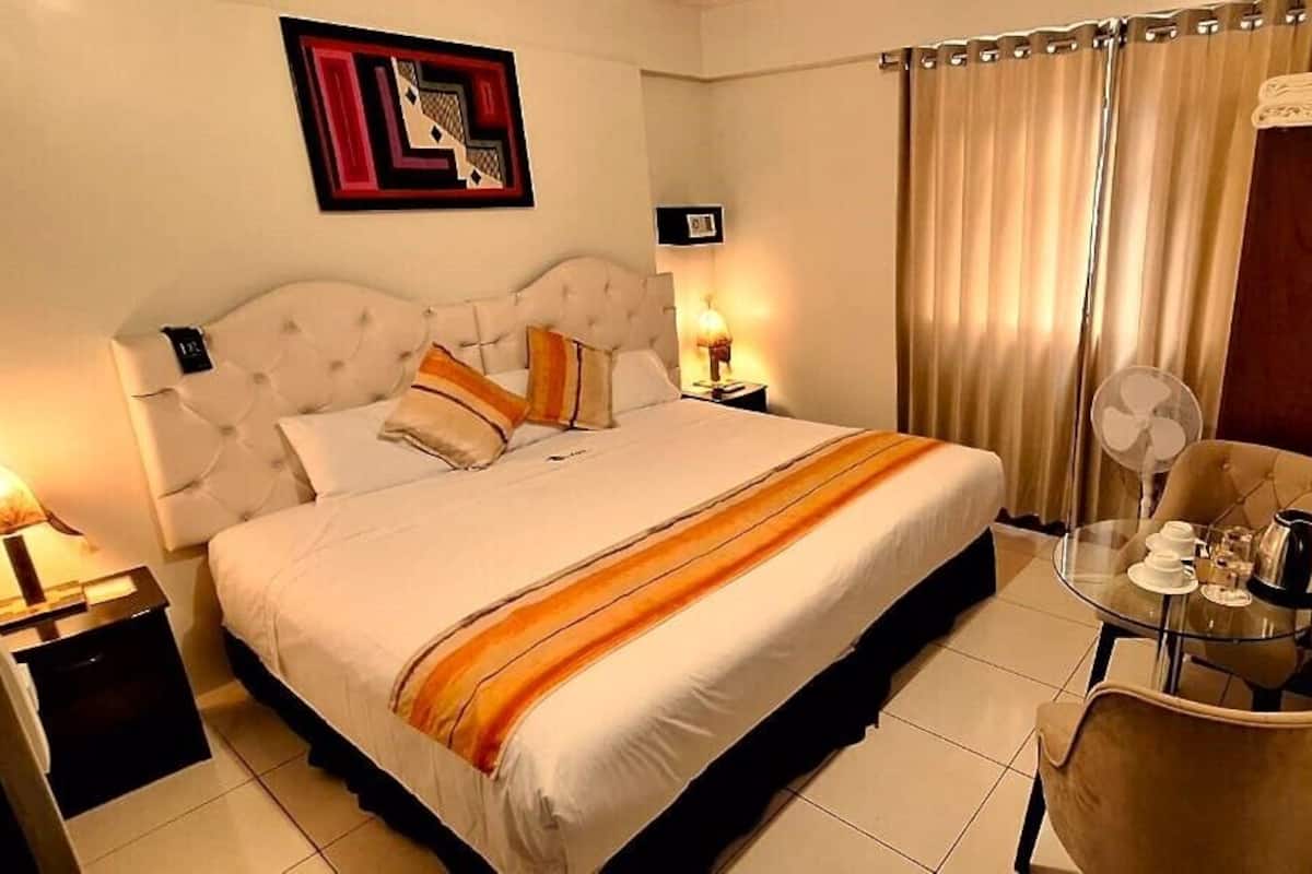 A hotel room with a large bed, a bedside table with a lamp, a chair, and a small table with a fan and a kettle.