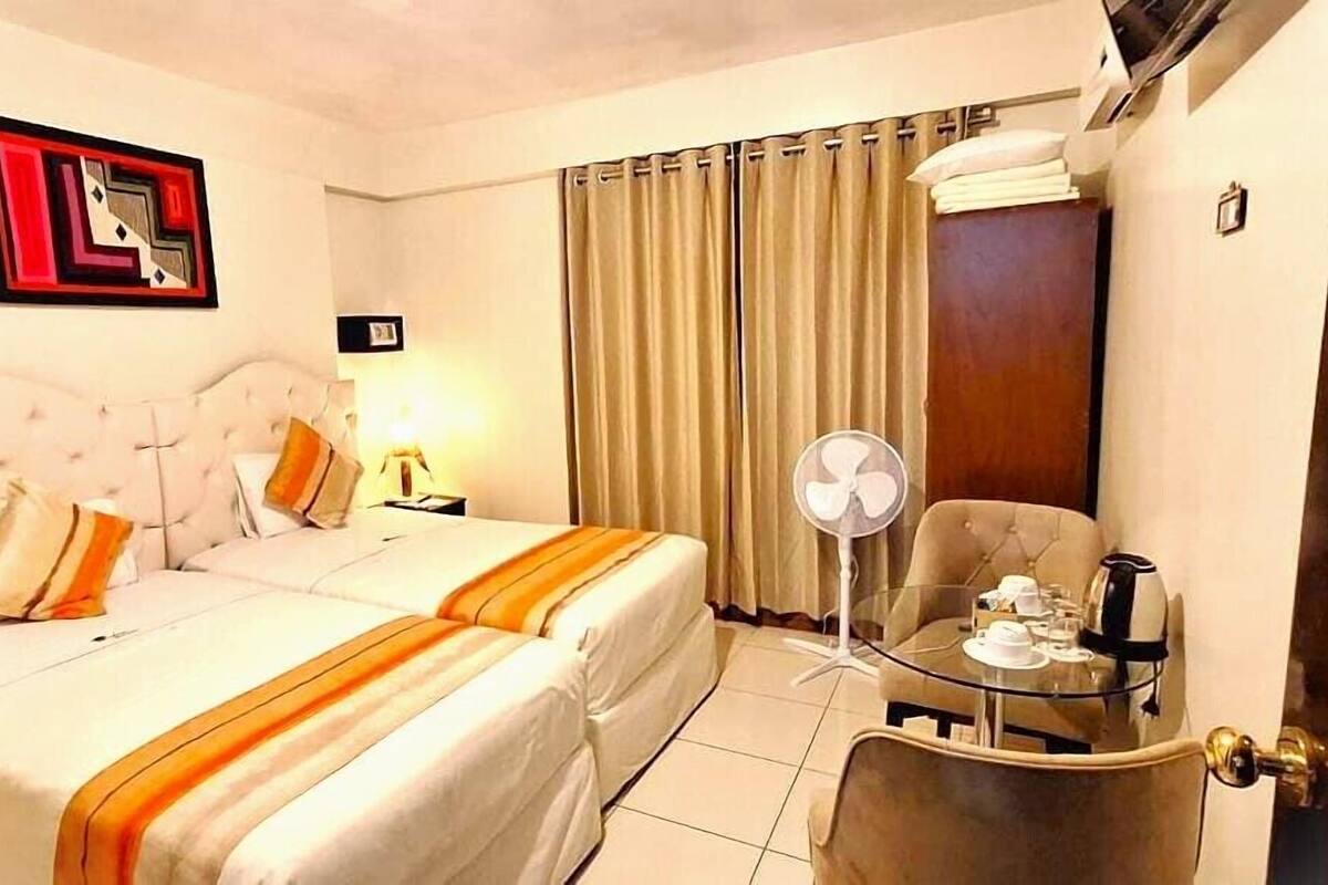 A hotel room with a bed, two bedside tables, a chair, a small table with a fan, and a picture on the wall.