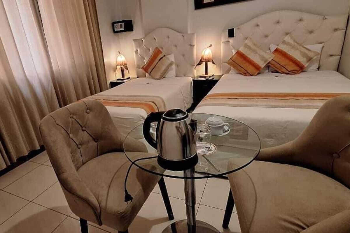 A hotel room with a bed, two bedside tables with lamps, a glass table with a coffee pot, and two armchairs.