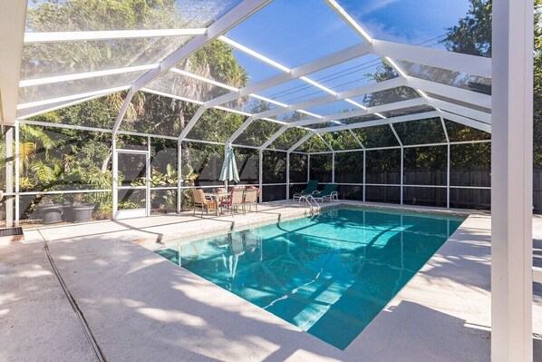 A covered outdoor swimming pool with a glass enclosure, surrounded by trees and a wooden fence.
