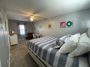 A bedroom with a bed, a desk, a chair, a ceiling fan, and a window with curtains.